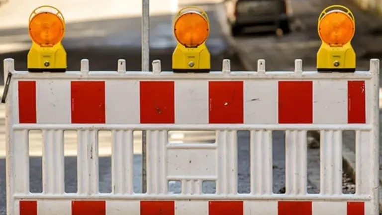 Ein rot-weiß gestreifter Bauzaun mit drei gelben blinkenden Warnleuchten auf einer städtischen Straße.