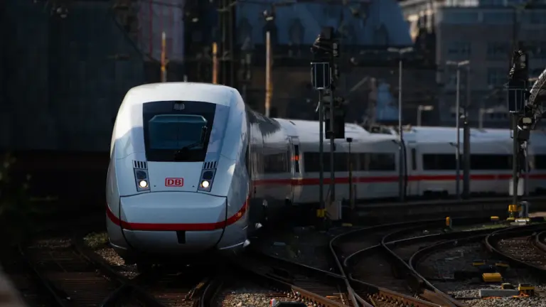 Ein weißer ICE-Zug fährt auf Gleisen an einem modernen Bahnhof in Deutschland entlang.
