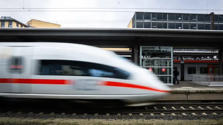 Ein weißer ICE-Zug fährt bei bewölktem Himmel durch einen deutschen Bahnhof.