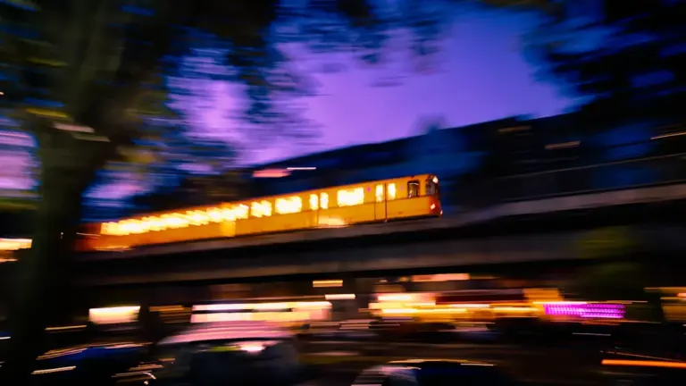 Gelbe Berliner U-Bahn fährt bei Dämmerung über eine Hochbahnstrecke mit Stadtlichtern im Hintergrund.