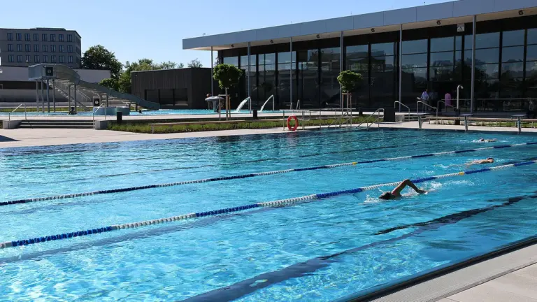 Ein hellblaues Außenbecken mit Schwimmern, Rutsche und modernem Glasgebäude an einem sonnigen Sommertag.