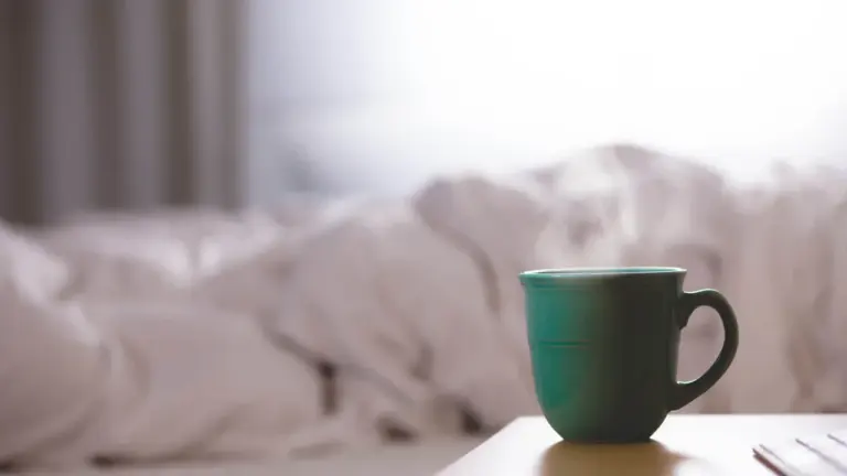 Eine dampfende Tasse auf einem Nachttisch vor einem ungemachten Bett am Morgen.