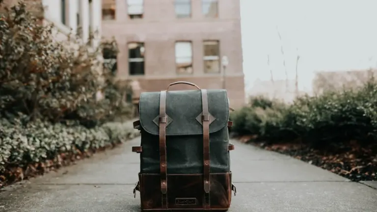 Ein grauer Rucksack steht auf einem Gehweg vor einem alten Schulgebäude in Deutschland.