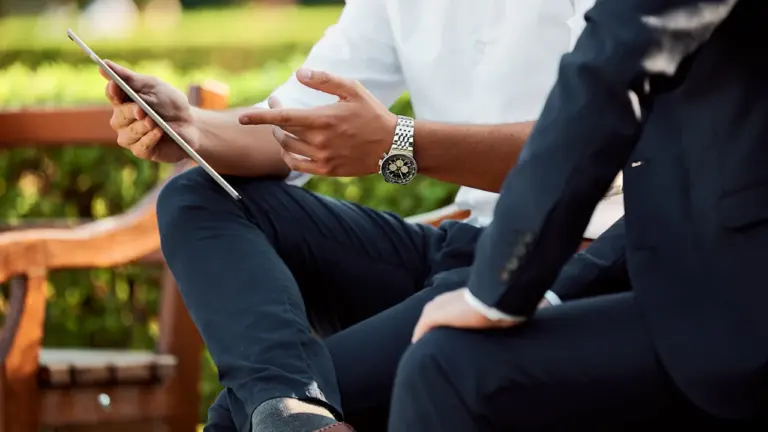 Zwei Personen im Business-Outfit sitzen auf einer Parkbank und besprechen wichtige Unterlagen.