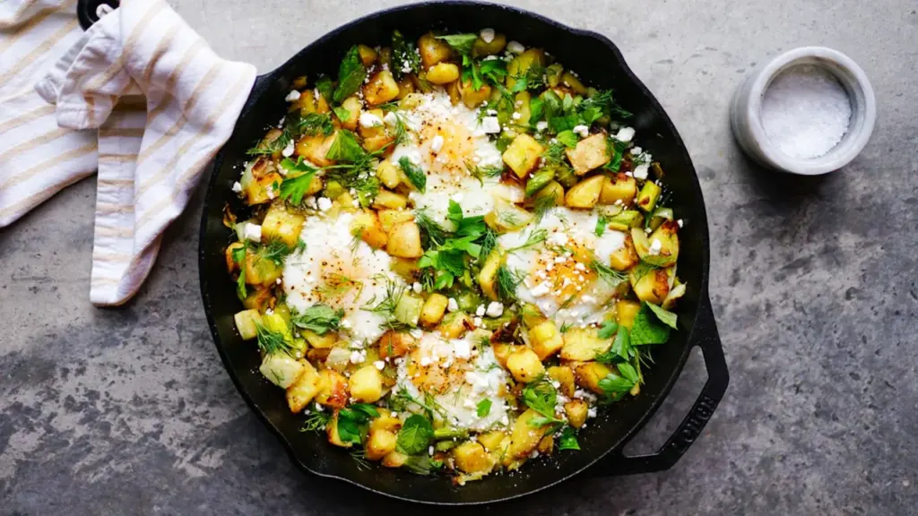 Eine gusseiserne Pfanne mit gebratenem Gemüse, Kirschtomaten und zerbröseltem Feta auf dunklem Untergrund.