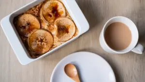 Ein Apfel-Hafer-Auflauf in einer weißen Auflaufform neben einer Tasse Kaffee auf Holztisch.