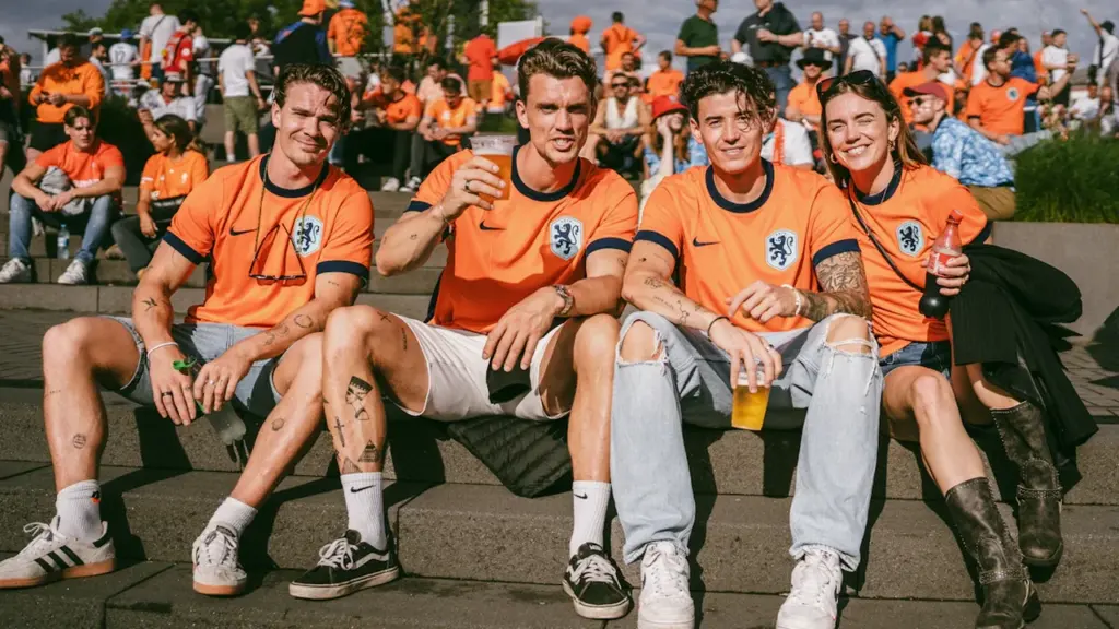Vier niederländische Fußballfans im Oranje-Trikot sitzen gemeinsam auf einer Treppe vor dem Stadion.