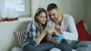 Junges Paar trinkt morgens Kaffee und schaut gemeinsam auf ein Smartphone im Bett.