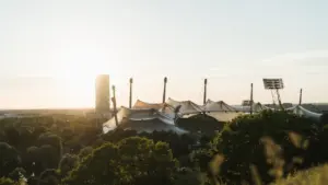 Das Münchner Olympiastadion eingebettet in eine grüne Parklandschaft bei sonnigem Wetter.